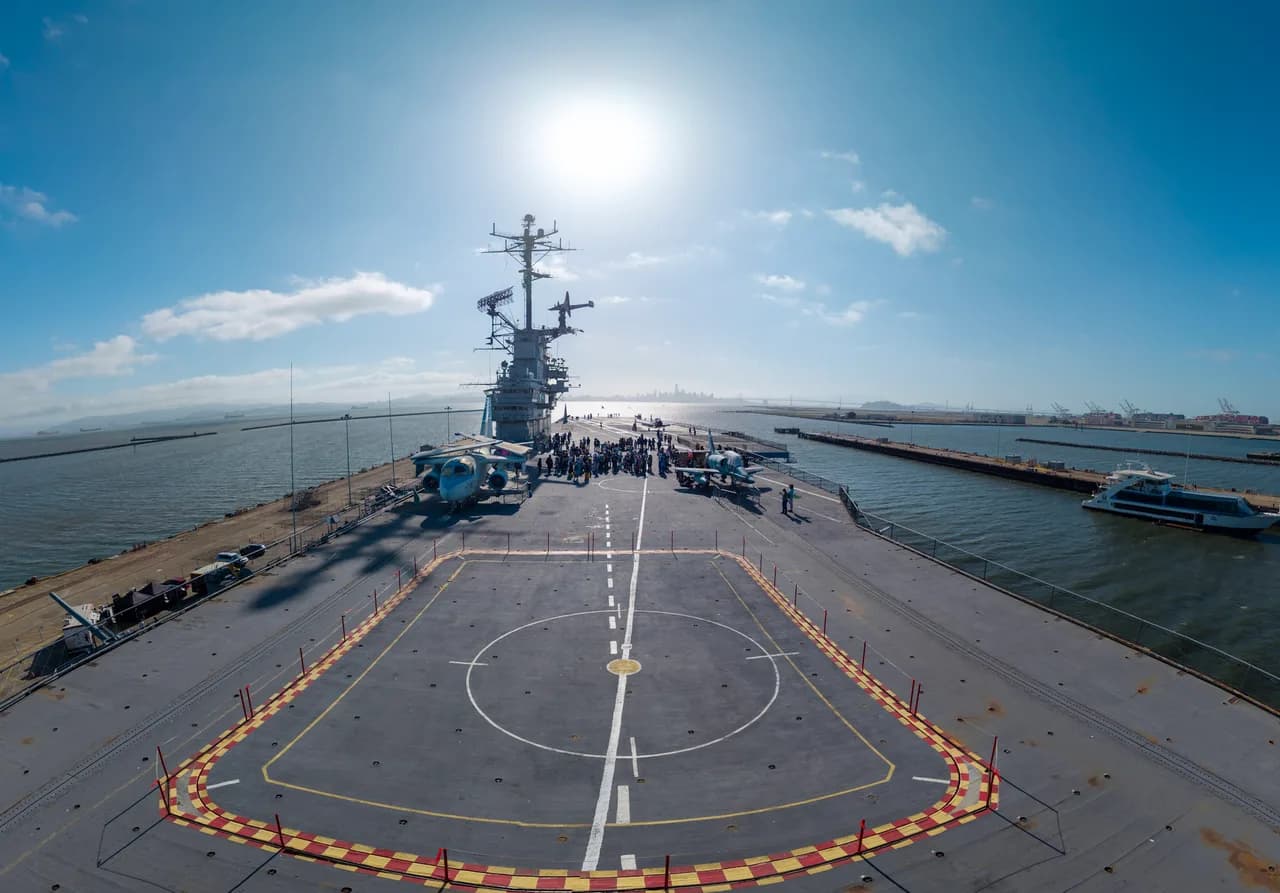 An aircraft carrier flight deck stretches toward its island tower, with sunlit water on both sides. Coast & City photo 6 of 11.