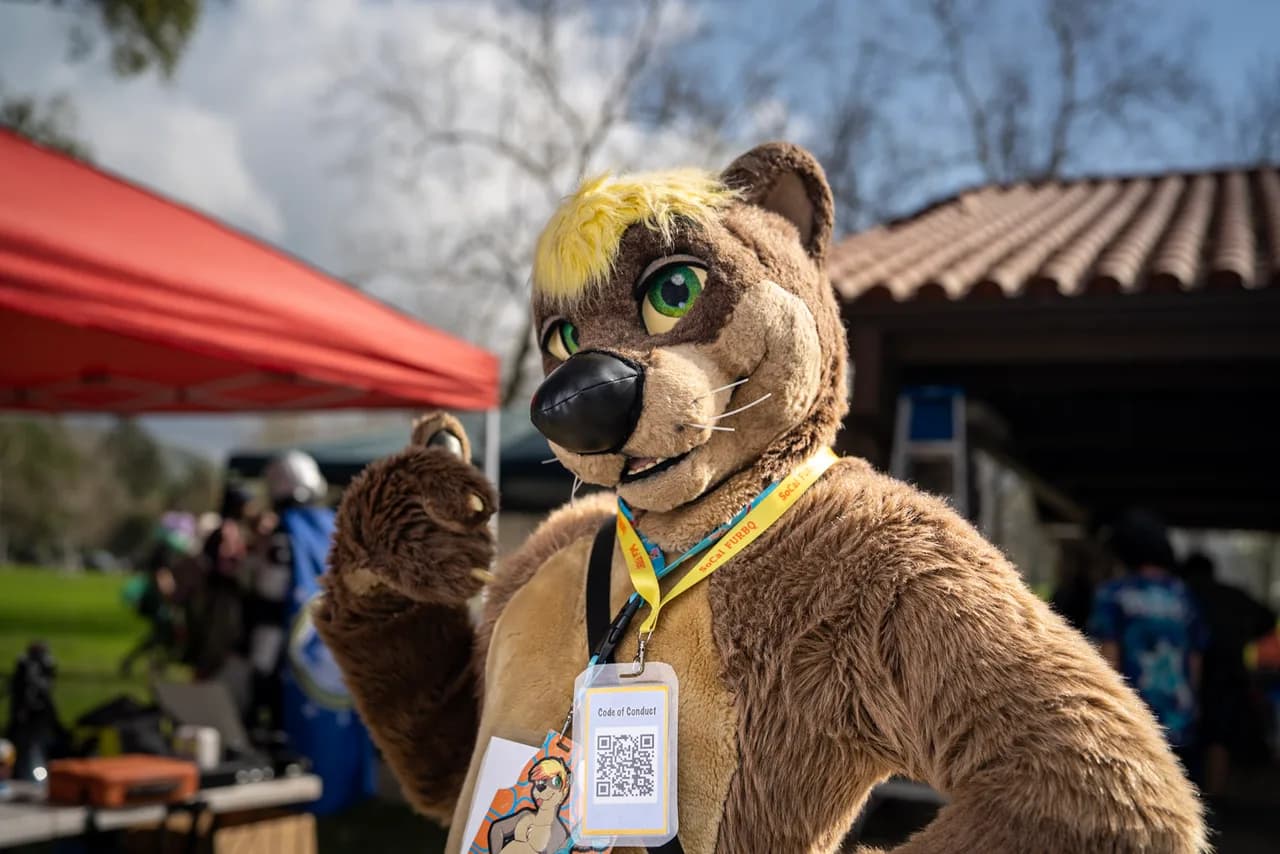 A person in a brown canine costume with green eyes and a yellow forelock raises one paw beside a pavilion. Costumes photo 6 of 20.