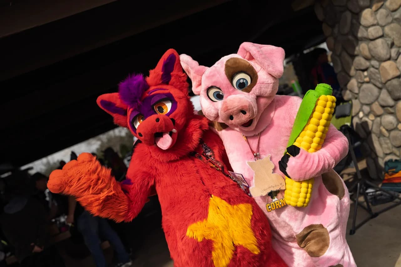 Two people in red and purple and pink animal costumes pose together, with one holding an oversized corn prop. Costumes photo 7 of 20.