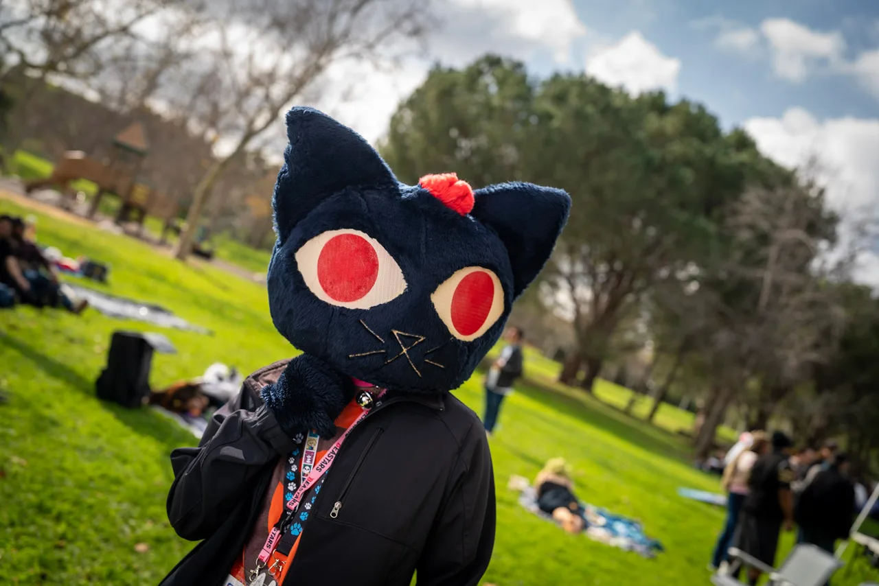 A person in a dark blue cat costume with large red eyes wears a black shirt in a sunny park. Costumes photo 9 of 20.
