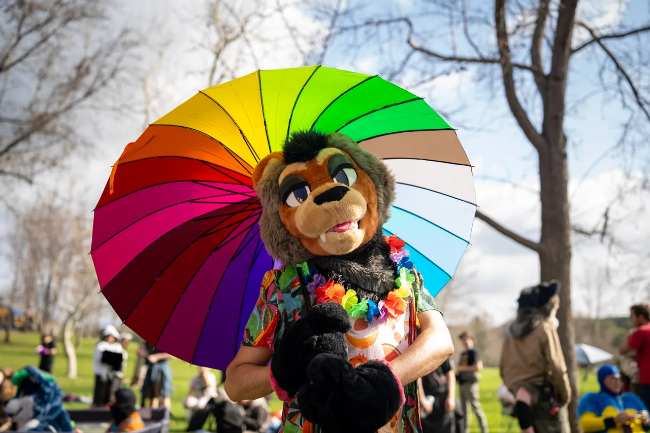 A person in a brown animal costume wears a flower lei while holding a rainbow umbrella in a park. Costumes photo 12 of 20.