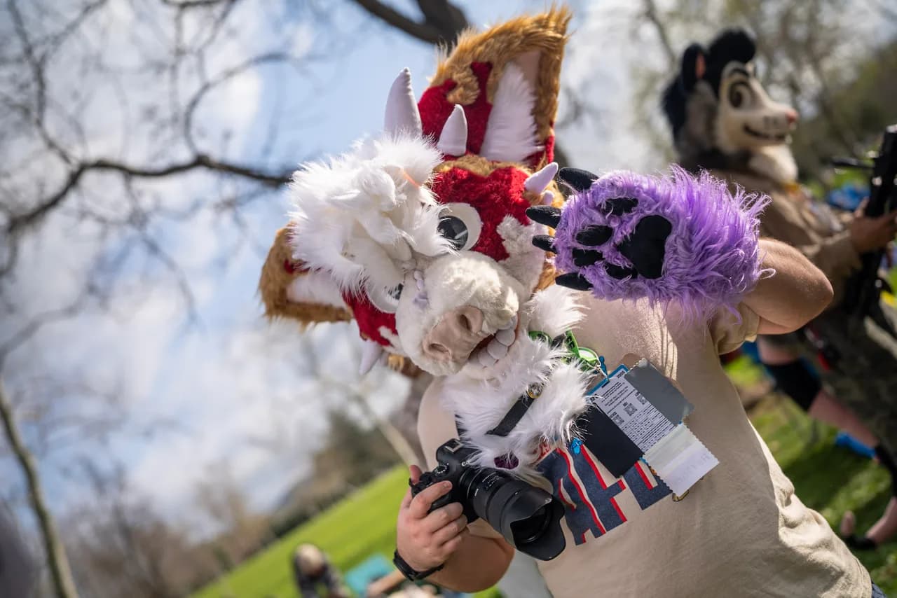 A person in a white and red animal costume raises a purple paw while holding a camera. Costumes photo 13 of 20.