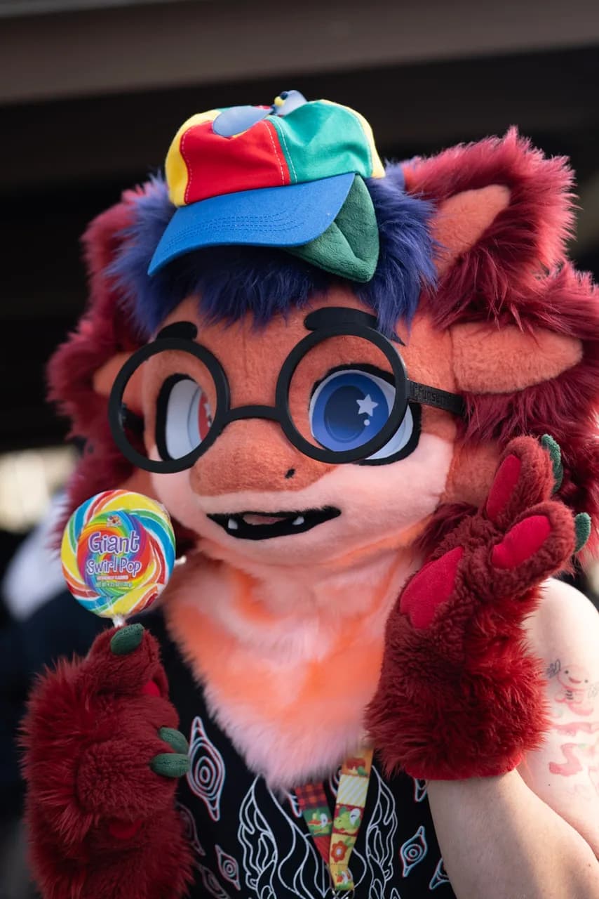 A person in a red and blue animal costume wears round glasses and a multicolored cap while holding a rainbow lollipop. Costumes photo 15 of 20.