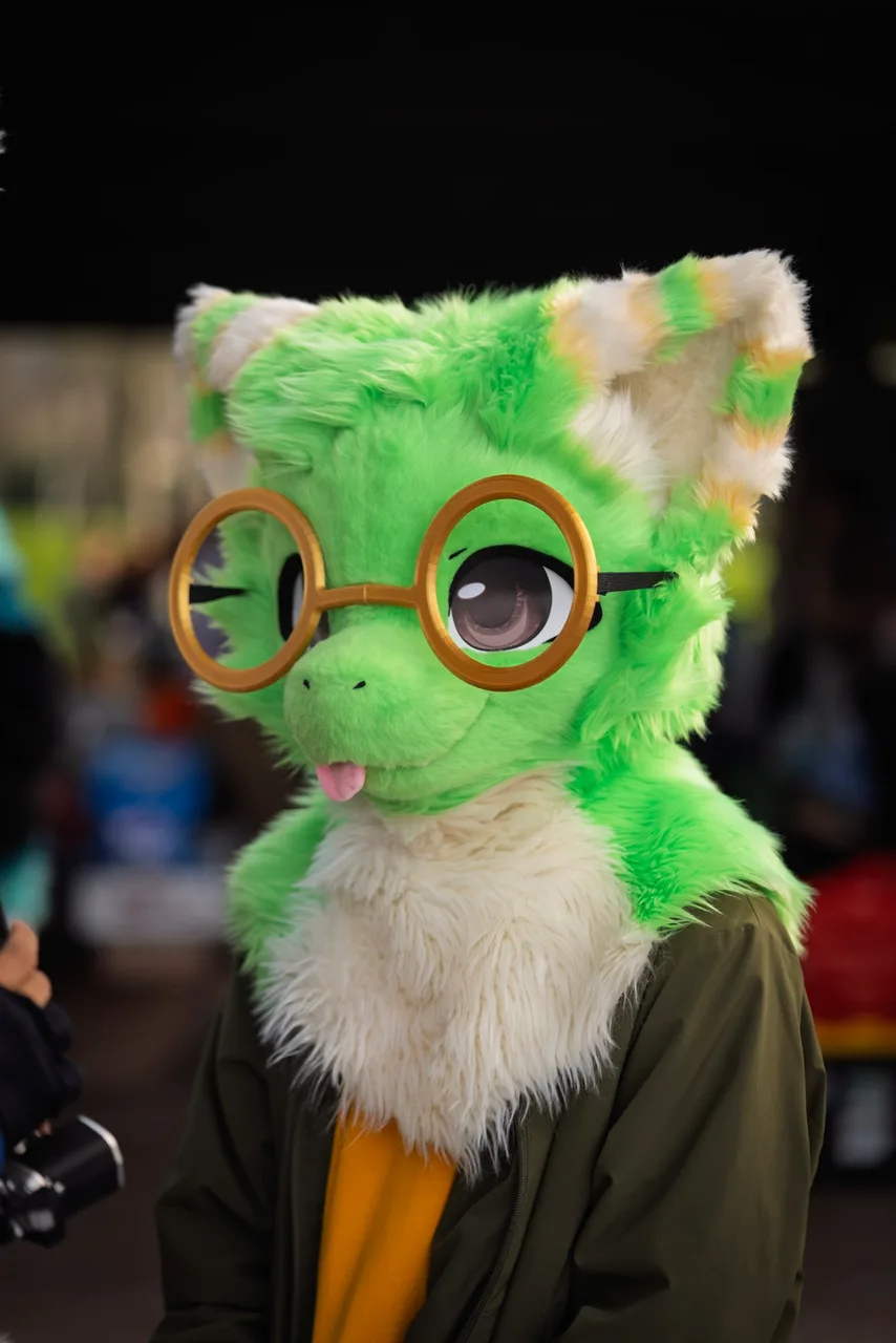 A person in a bright green cat costume wears round gold glasses and a dark jacket. Costumes photo 16 of 20.