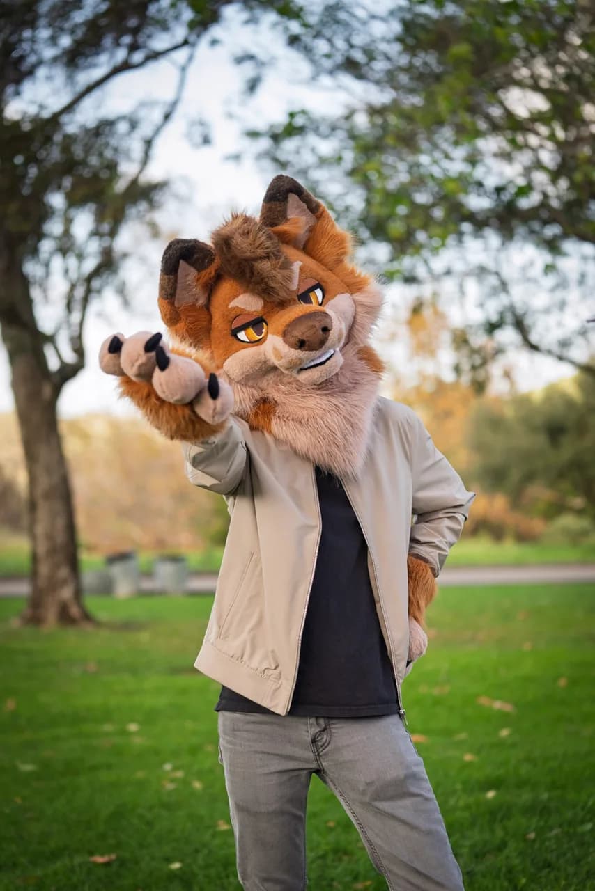 A person in a brown canine costume and a light jacket reaches one paw toward the camera in a park. Costumes photo 17 of 20.