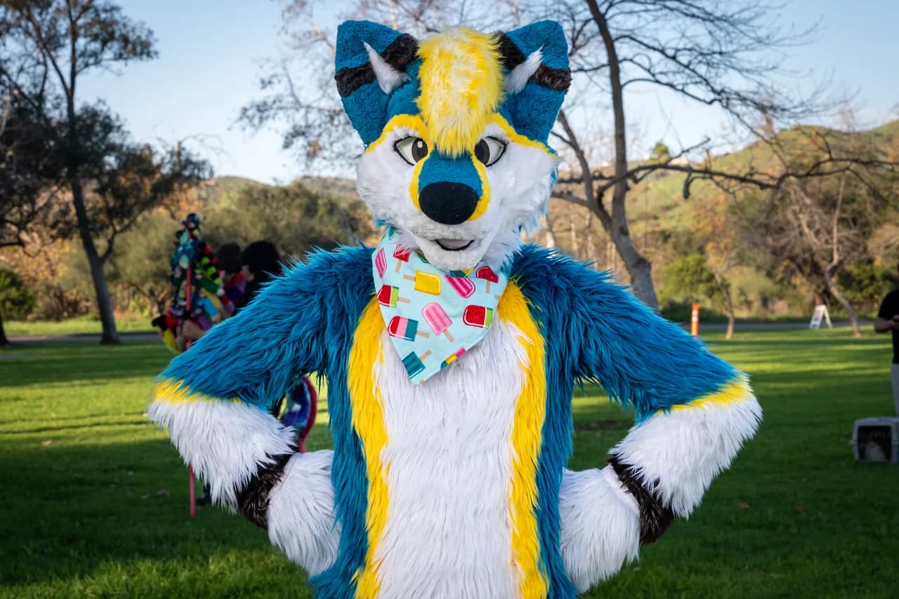 A person in a blue, yellow, and white canine costume poses with hands on hips and a colorful bandana. Costumes photo 18 of 20.