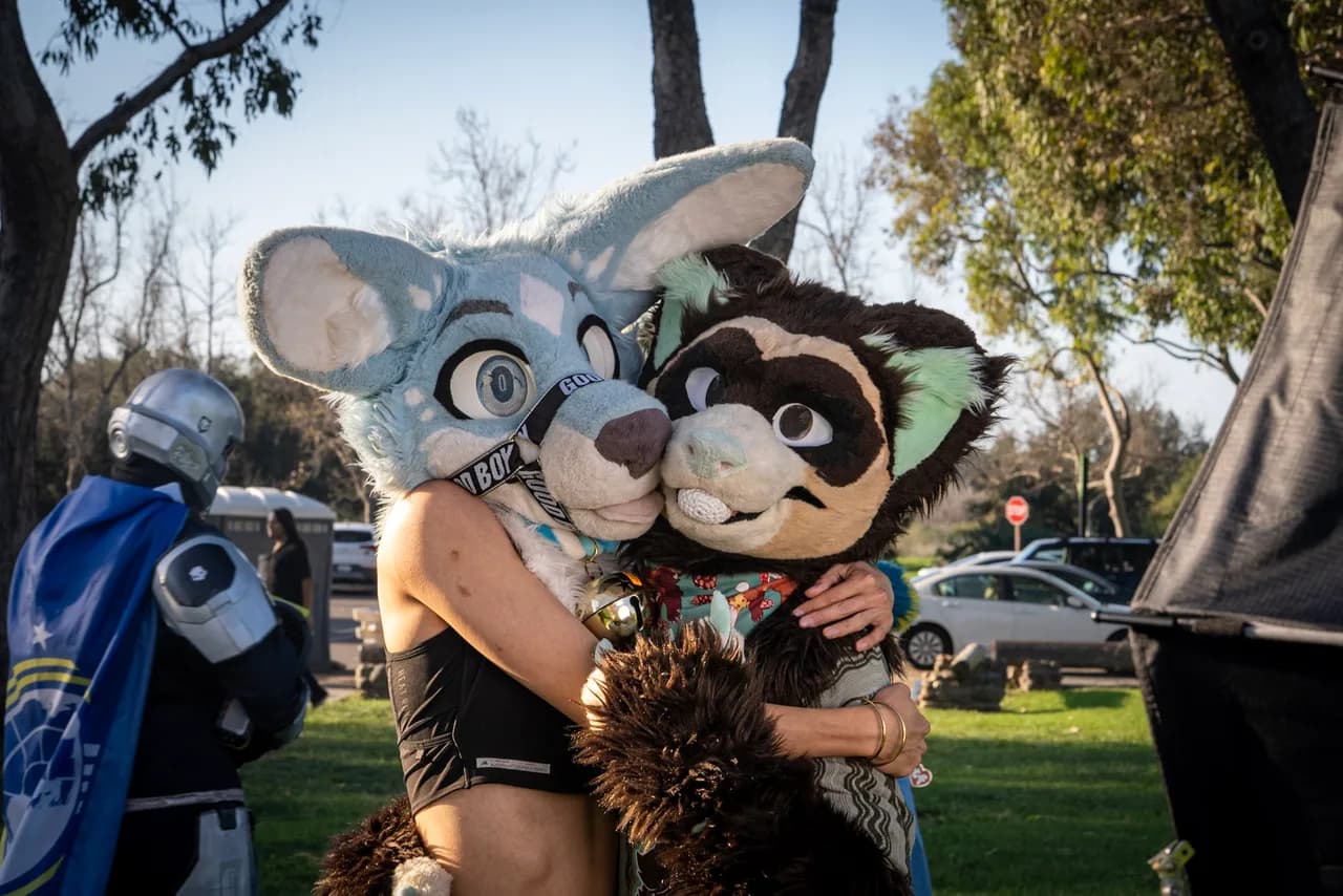 Two people wearing a gray rabbit head and a brown animal head hug beneath trees at an outdoor gathering. Costumes photo 19 of 20.